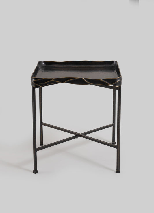 A black metal side table with a rectangular tray top featuring a slightly wavy, textured edge. The table has a minimalist frame with crossed supports and small square feet, emphasizing modern and industrial design.