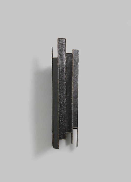 A modern wall-mounted sculpture made of blackened metal with a textured, rough surface and vertical rectangular panels. The piece has an abstract, minimalist design with clean lines and a dark matte finish.