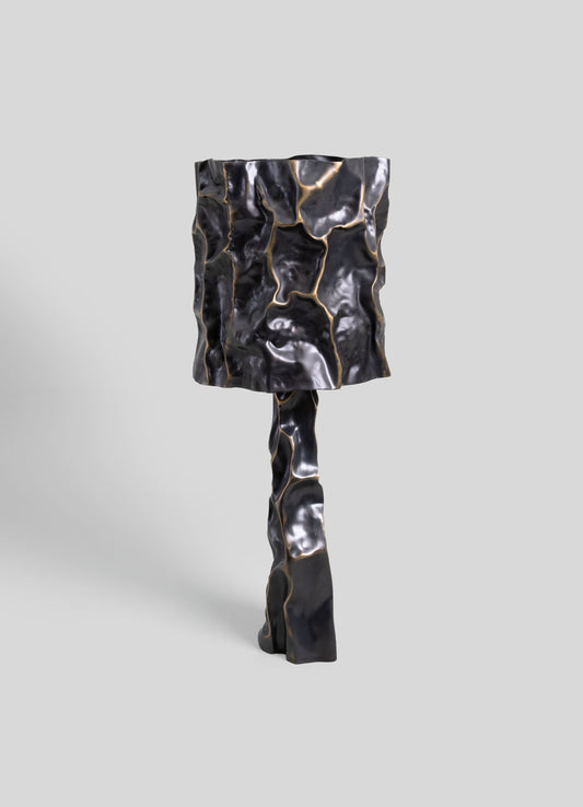A sculptural table lamp with a textured, abstract design in black and metallic gold accents, featuring a standing base and a rectangular lampshade with a crumpled, organic surface.