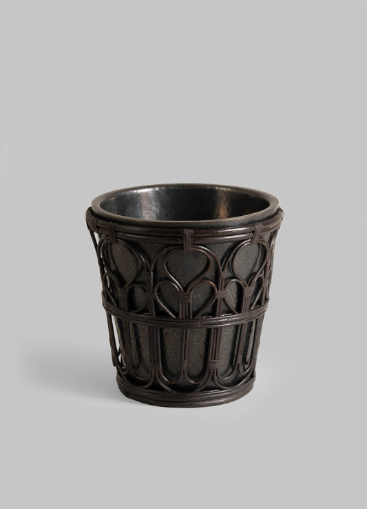 The VIYA Manipur Champagne Bucket is a dark, round container featuring intricate woven patterns and curved designs—perfect for elegant home decor. Displayed against a plain gray background.