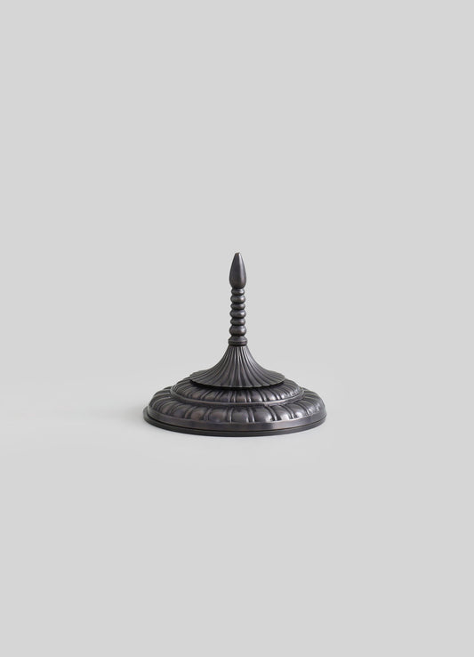A dark metal candle holder with an intricate, tiered design and a pointed finial on top, featuring a textured, rounded base.