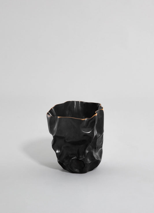 A black ceramic vase with an irregular, textured surface and wavy rim, set against a plain light gray background.