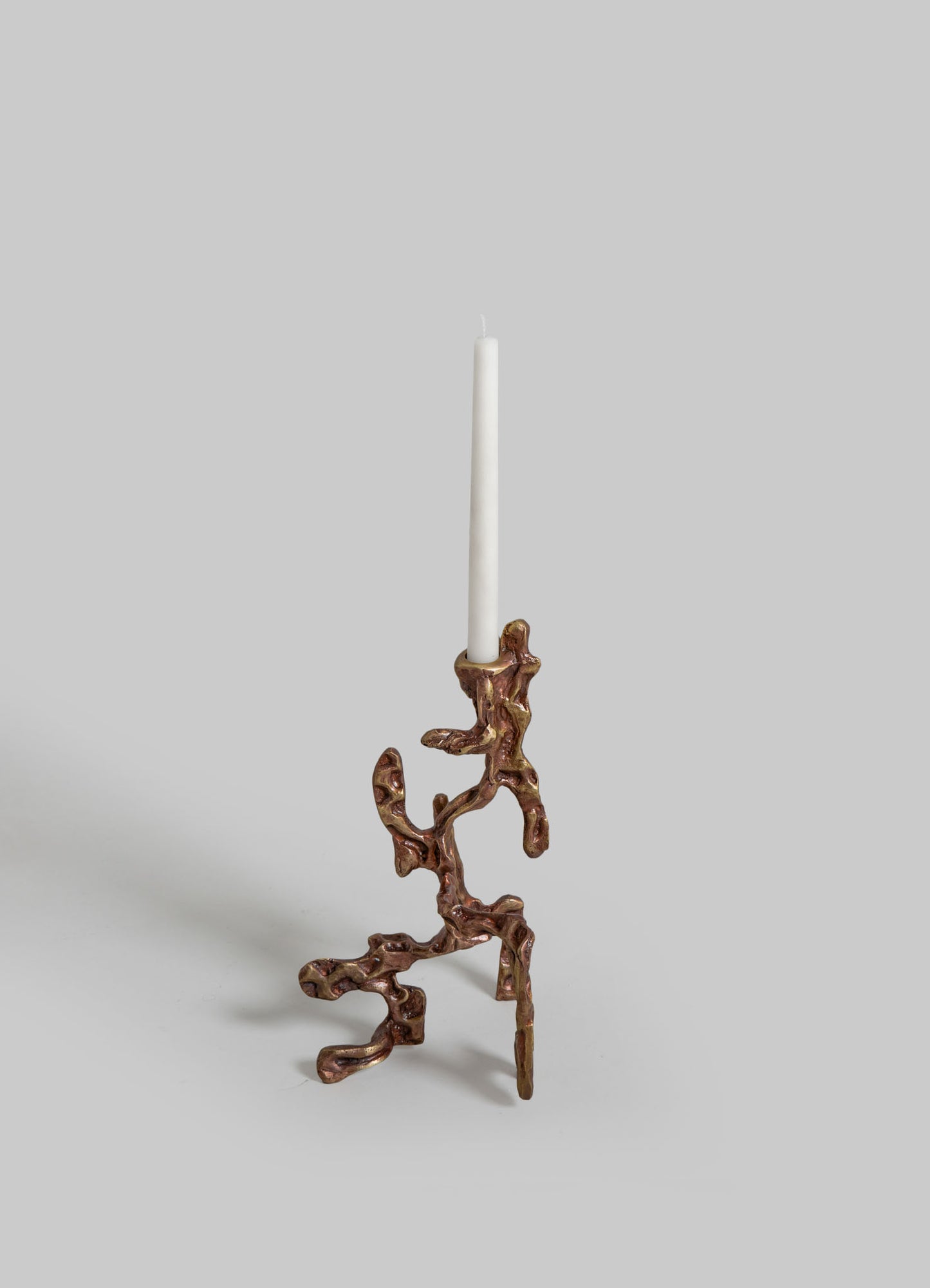 A sculptural candlestick holder made of metal with an organic, twisted design and a bronze finish, holding a tall white taper candle. The piece has a contemporary, handcrafted style with a naturalistic, abstract motif.