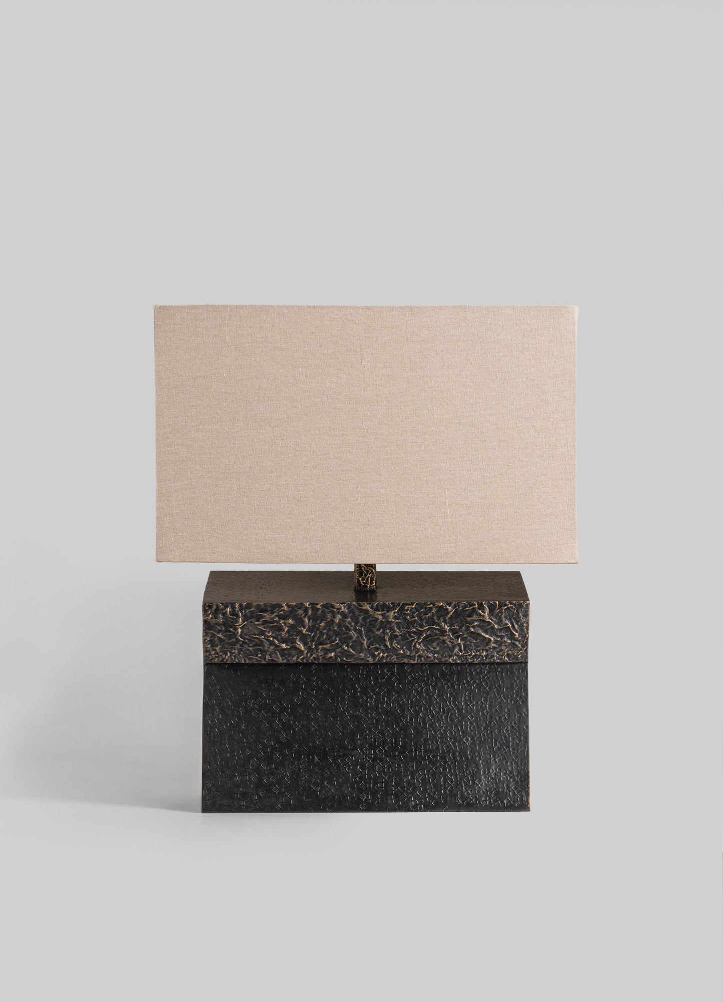 Table lamp with a rectangular fabric shade in neutral beige tones, mounted on a base with a textured black and dark brown finish. The design is contemporary with a minimalist silhouette and contrasting materials.