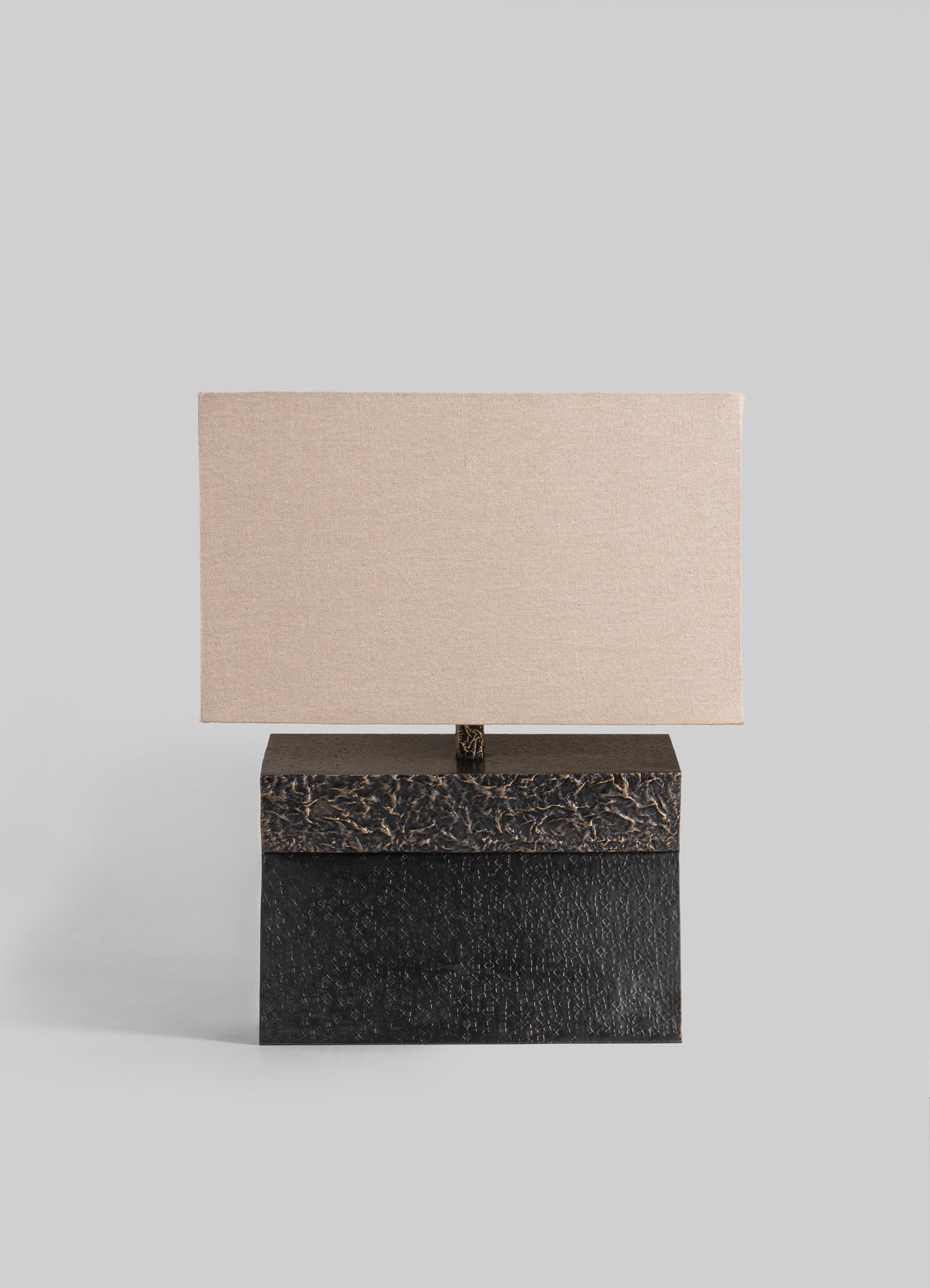 Table lamp with a rectangular fabric shade in neutral beige tones, mounted on a base with a textured black and dark brown finish. The design is contemporary with a minimalist silhouette and contrasting materials.