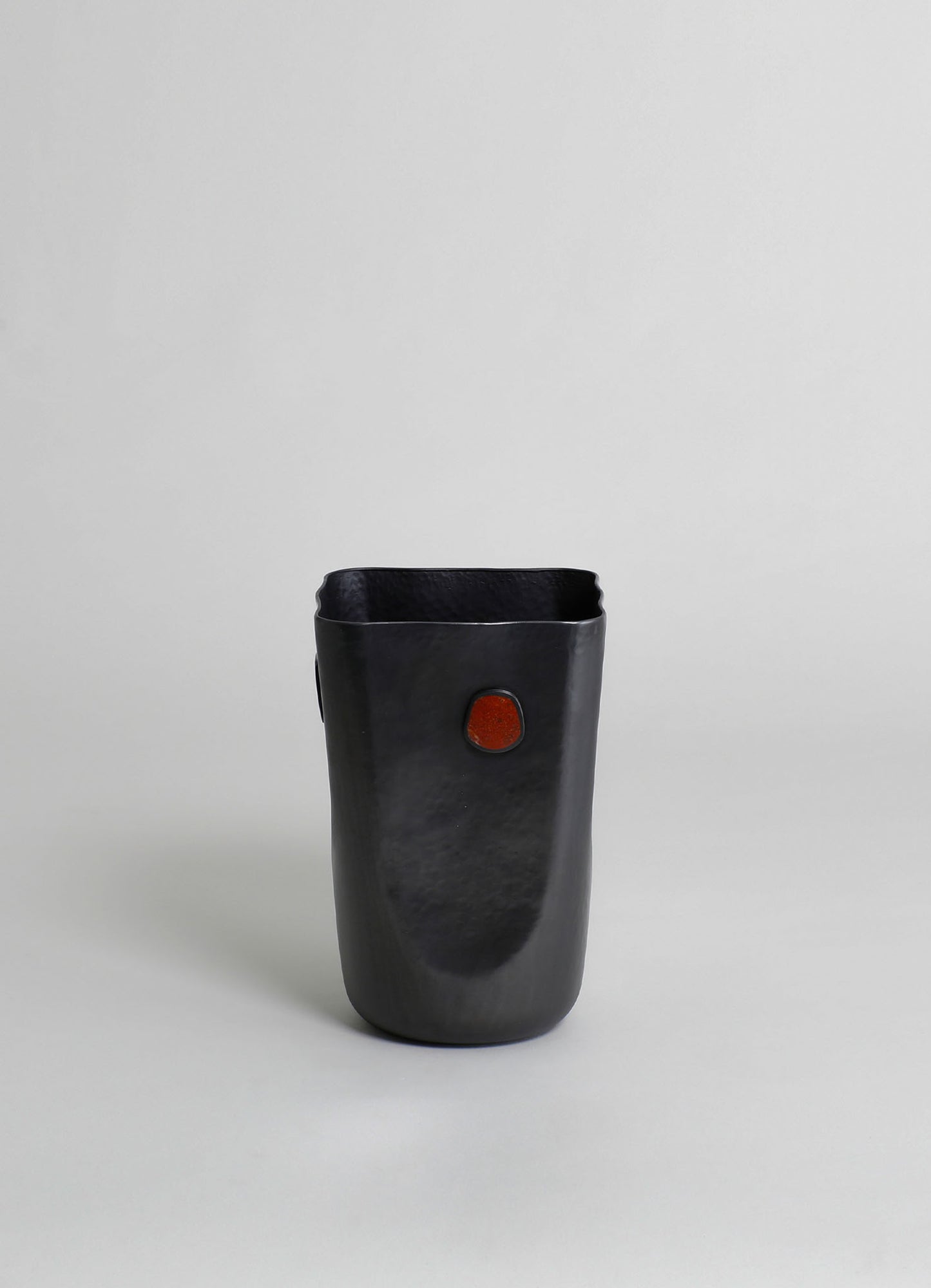 A black ceramic vase with a matte finish and a small red accent on the front, featuring an irregular, organic shape and smooth surface.