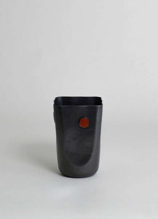 A black ceramic vase with a matte finish and a small red accent on the front, featuring an irregular, organic shape and smooth surface.