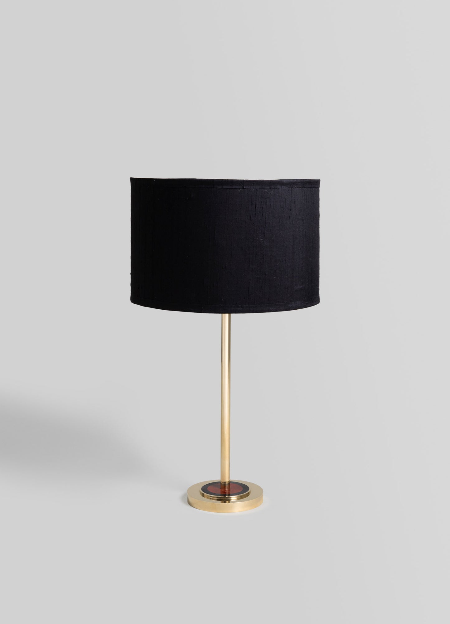 A table lamp with a cylindrical black fabric lampshade and a slim, metallic gold base on a round platform. The design is modern and minimalist with a contemporary style.