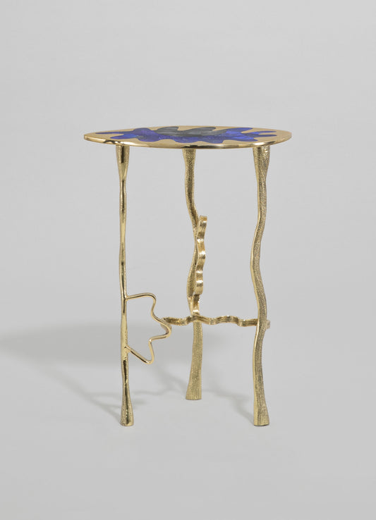 A side table with a brass finish and a sculptural, irregular form, featuring a round top with a painted floral motif in shades of blue and gray. The table has a contemporary style with a textured metallic surface and wavy, organic legs.