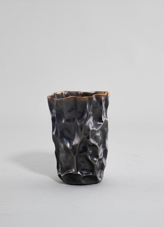 A decorative ceramic vase with an irregular, organic shape and a textured, glossy black finish with subtle metallic accents, set against a plain light background.