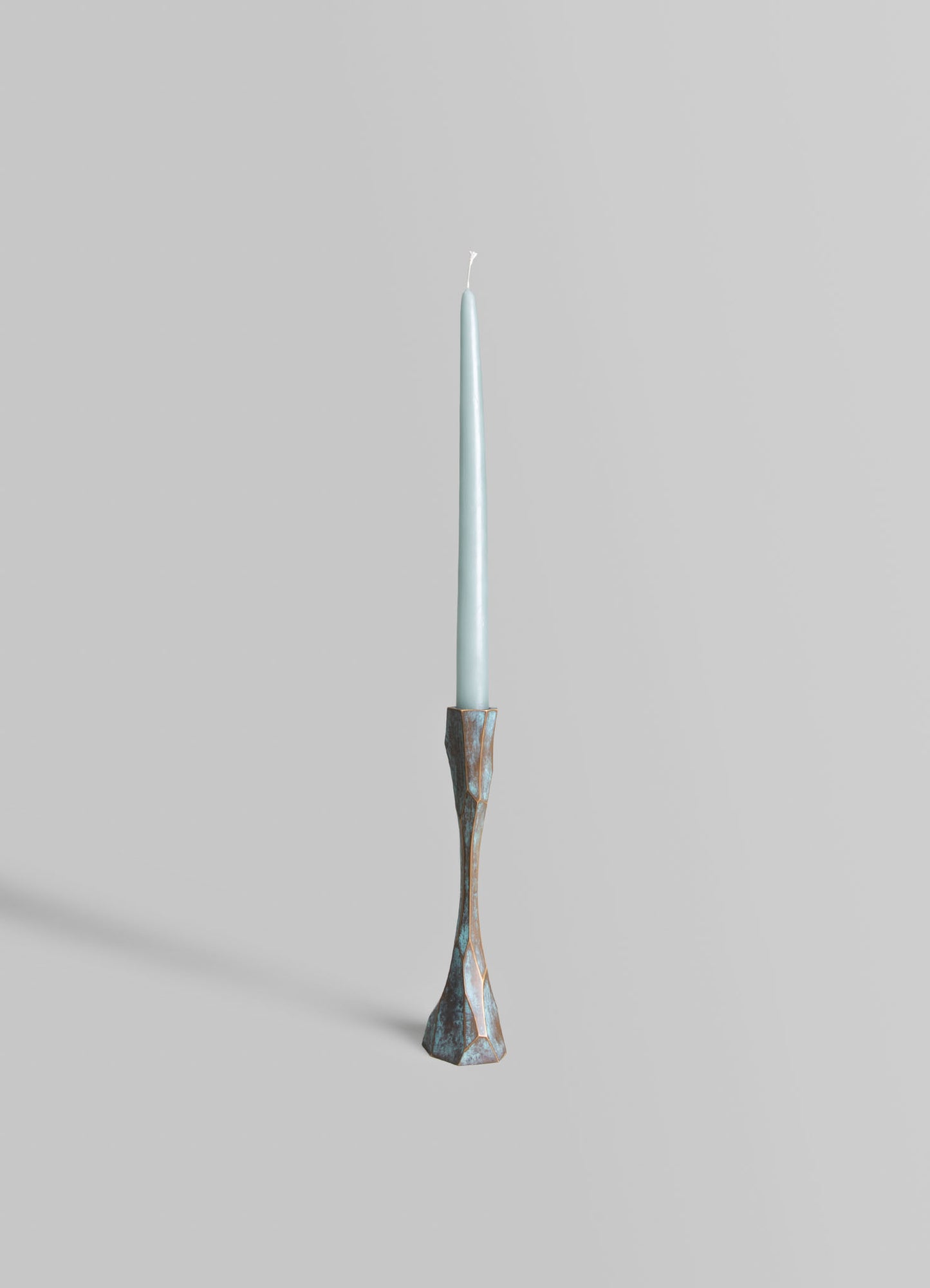 A tall, slender candle with a light blue color and a wick at the top, mounted on a decorative holder with an irregular, geometric shape and a weathered silver finish.