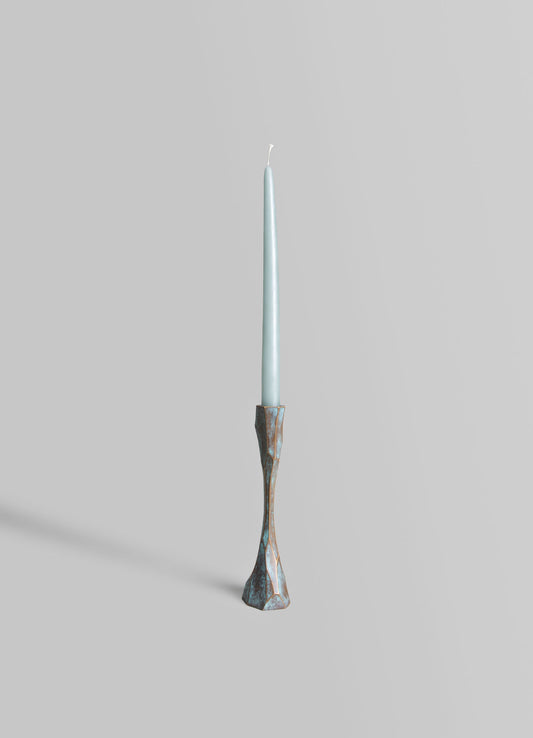 A tall, slender candle with a light blue color and a wick at the top, mounted on a decorative holder with an irregular, geometric shape and a weathered silver finish.