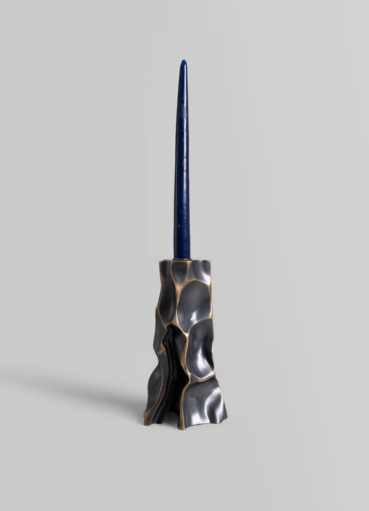 A sculptural candle holder with an abstract, organic shape in matte black and bronze tones, holding a tall, slender dark blue candle.