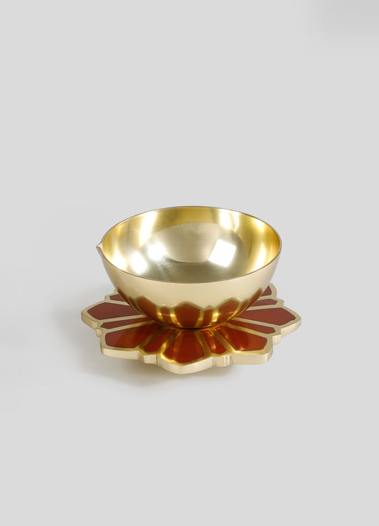 A metallic gold bowl with a reflective finish sits on a decorative, patterned base in red and gold. The bowl has a smooth, rounded shape, and the base features a petal-like design with bold, contrasting colors.