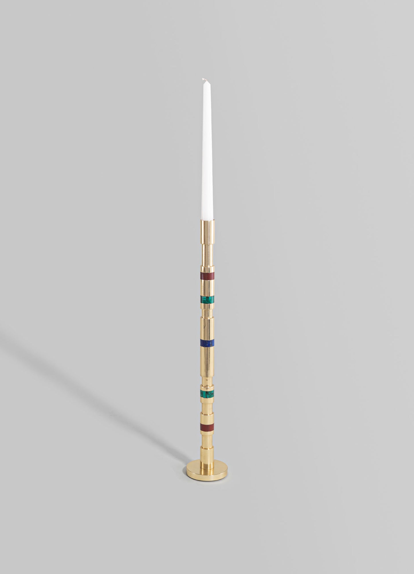 A tall candle holder with a slender, cylindrical brass body featuring colorful ring accents in red, green, and blue, mounted on a round brass base. The design has a modern and minimalist style with vibrant detailing.