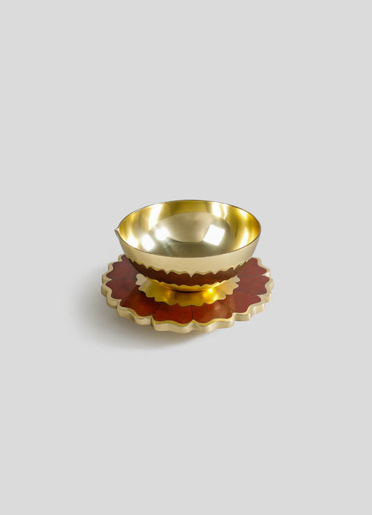 A brass bowl with a polished, reflective finish sits on a decorative ceramic coaster with a scalloped edge and a reddish-brown floral motif.