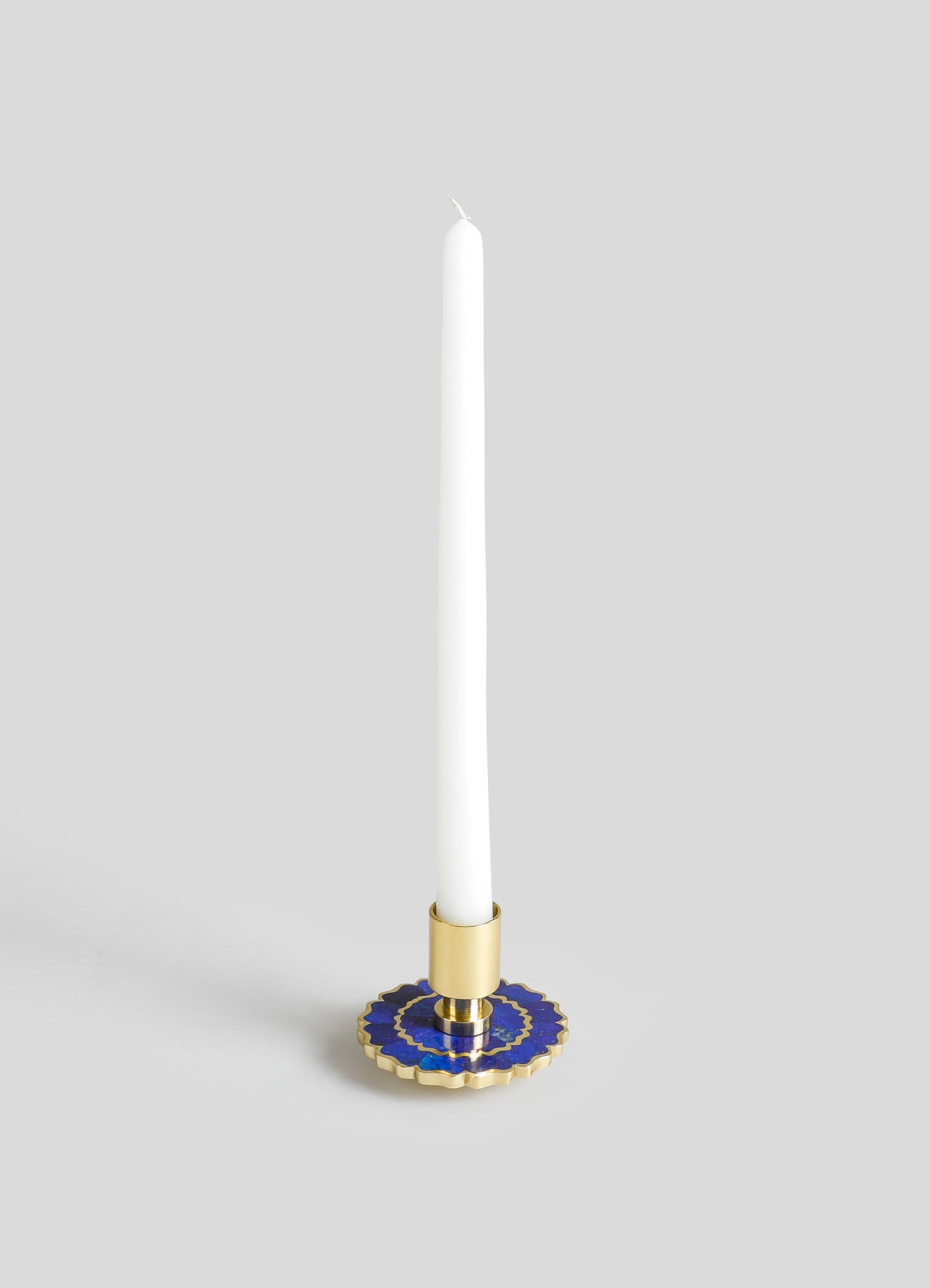 A candlestick holder made of gold-toned metal with a circular base featuring blue and gold decorative motifs. It holds a tall, white taper candle.