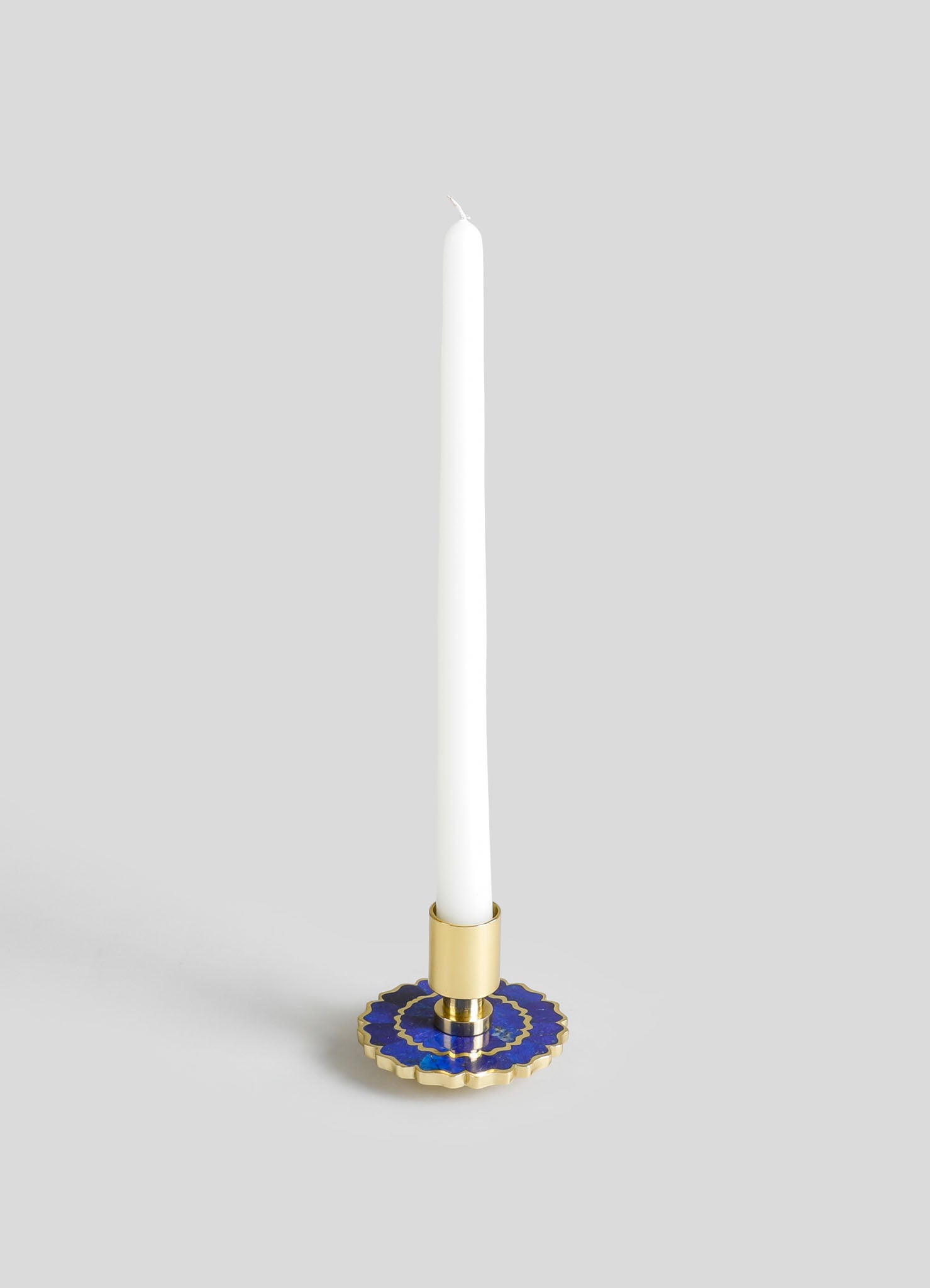 A candlestick holder made of gold-toned metal with a circular base featuring blue and gold decorative motifs. It holds a tall, white taper candle.