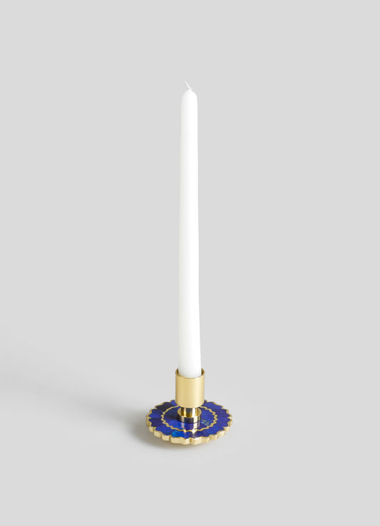 A candlestick holder made of gold-toned metal with a circular base featuring blue and gold decorative motifs. It holds a tall, white taper candle.