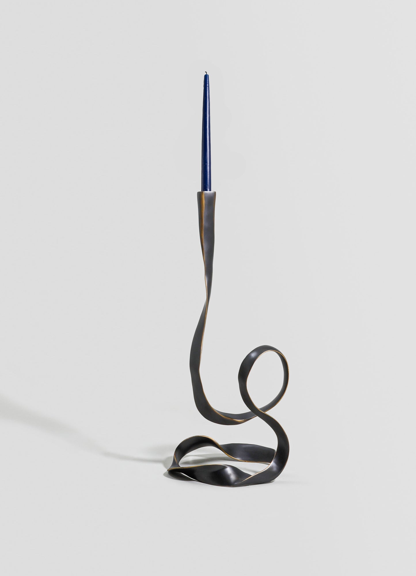 A sculptural candle holder made of metal with a twisted, flowing design in dark matte finish, holding a tall, slender blue candle. The piece has an abstract, contemporary style with an artistic, minimalistic aesthetic.