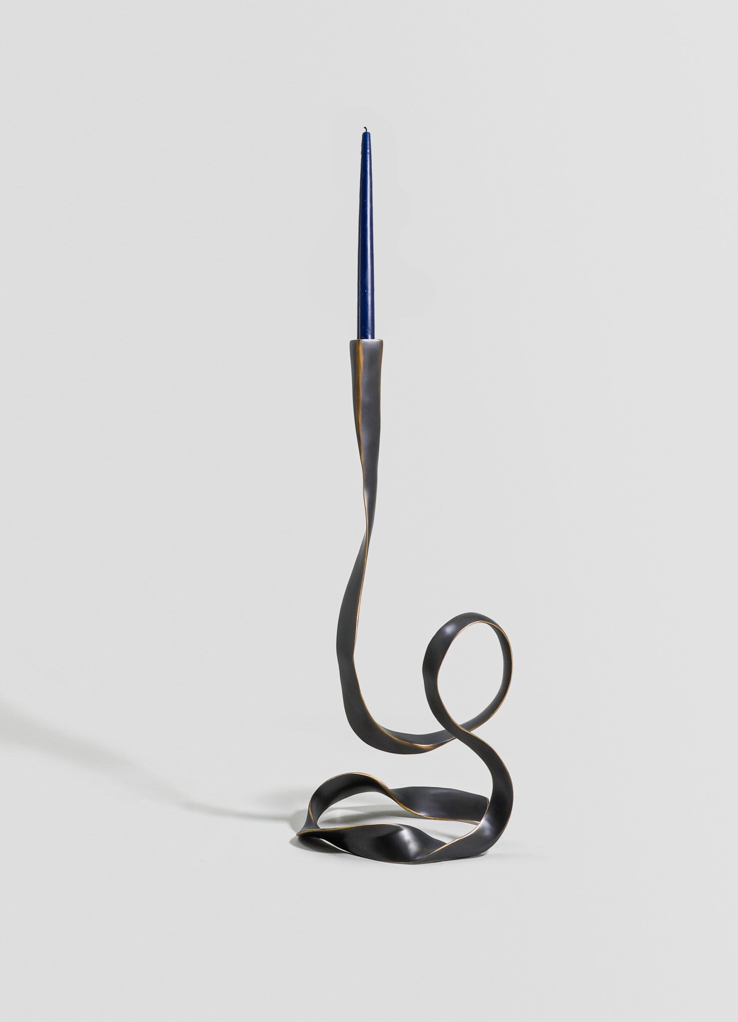 A sculptural candle holder made of metal with a twisted, flowing design in dark matte finish, holding a tall, slender blue candle. The piece has an abstract, contemporary style with an artistic, minimalistic aesthetic.