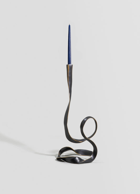 A sculptural candle holder made of metal with a twisted, flowing design in dark matte finish, holding a tall, slender blue candle. The piece has an abstract, contemporary style with an artistic, minimalistic aesthetic.