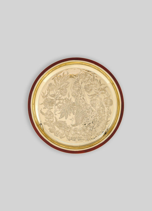 A round decorative tray with a metallic gold surface and intricate engraved motifs featuring a peacock, floral patterns, and foliage, bordered by a dark wooden rim.