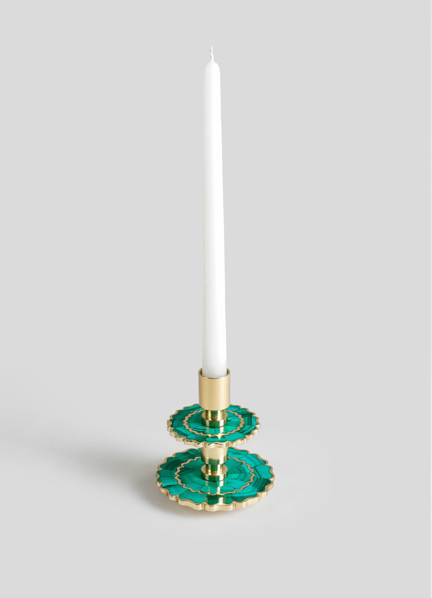 A tall white candle on a decorative candlestick holder with layered turquoise and gold accents, made of metal or enamel with scalloped edges. The overall style is elegant and contemporary, combining vibrant colors with metallic finishes.
