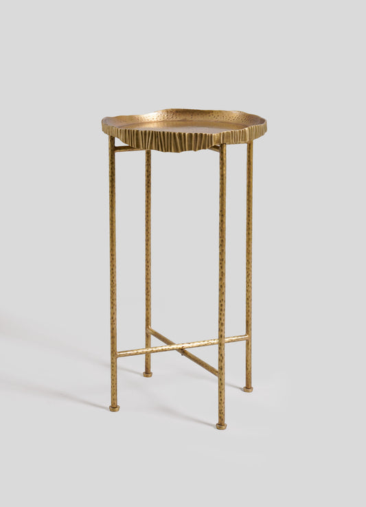 A tall metal side table with a hammered gold finish, featuring a scalloped edge tabletop and a minimalist frame with slender, dotted legs.