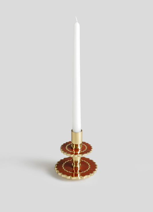 A tall white taper candle in a decorative holder with a gold and red geometric and floral design, featuring scalloped edges and a glossy finish.