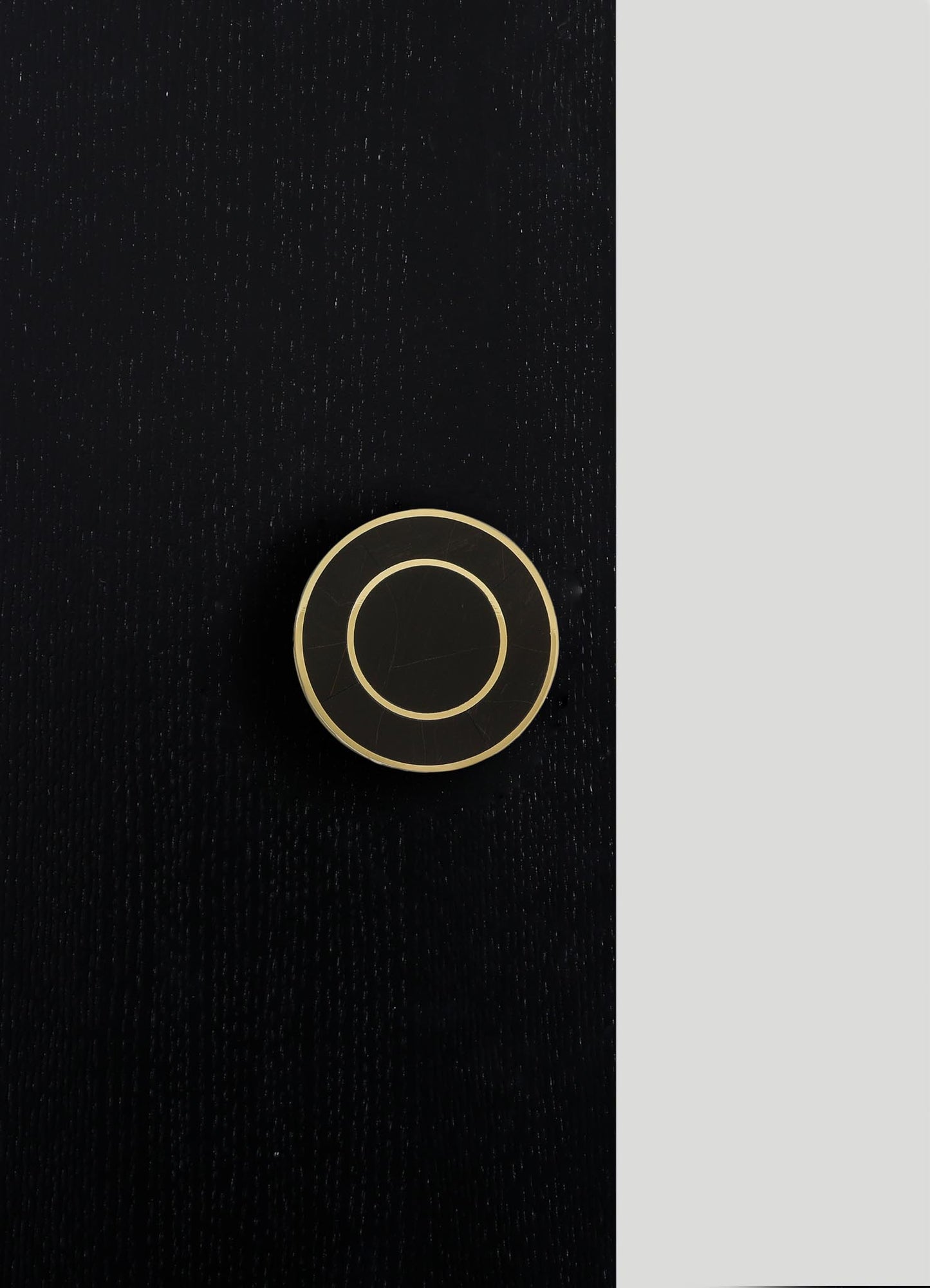A round, black ceramic object with concentric beige rings and a glossy finish, placed on a textured black surface with a vertical light gray background.