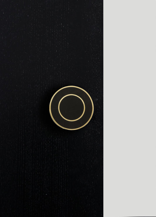 A round, black ceramic object with concentric beige rings and a glossy finish, placed on a textured black surface with a vertical light gray background.