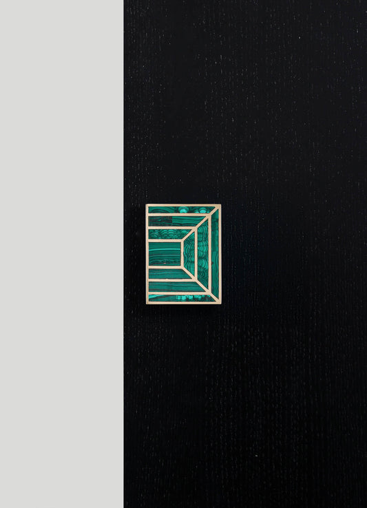 A geometric brooch with a rectangular shape, featuring layered, concentric lines in shades of teal and turquoise, mounted on a black textured surface.