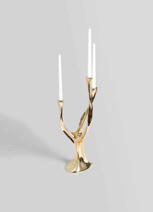 A decorative candle holder made of polished brass with an organic, twisting design resembling intertwined branches. It holds three tall white taper candles and has a contemporary, luxurious style.