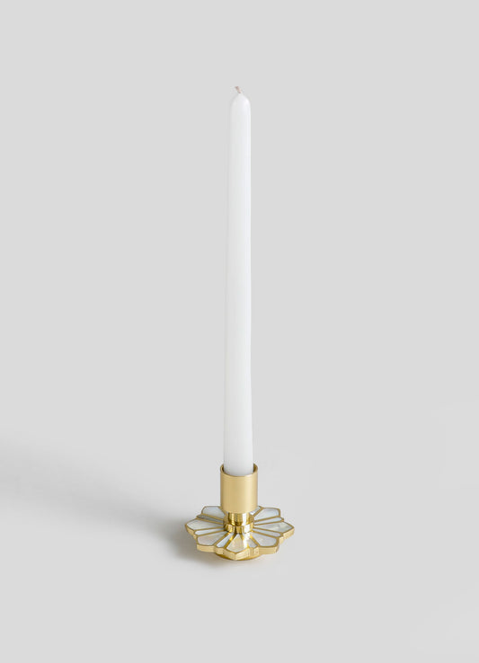 A tall, slim candle with a white body and unburned wick, placed in a decorative gold and white holder with a geometric, floral-inspired pattern.