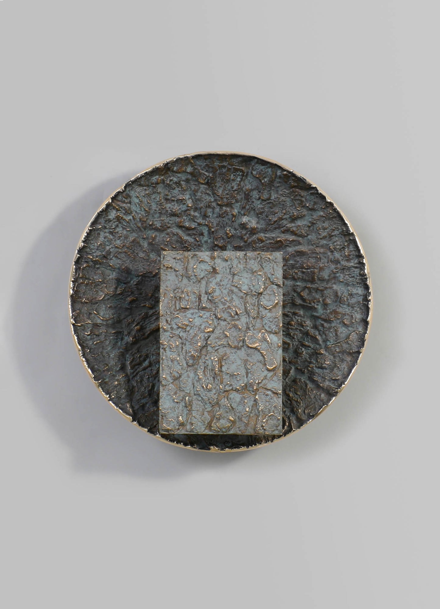 A round, textured metal plate with a distressed finish in dark and tarnished tones, featuring a rectangular inset with a similar textured pattern and metallic accents.