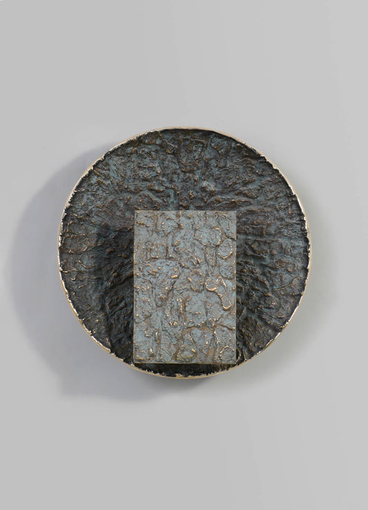 A round, textured metal plate with a distressed finish in dark and tarnished tones, featuring a rectangular inset with a similar textured pattern and metallic accents.