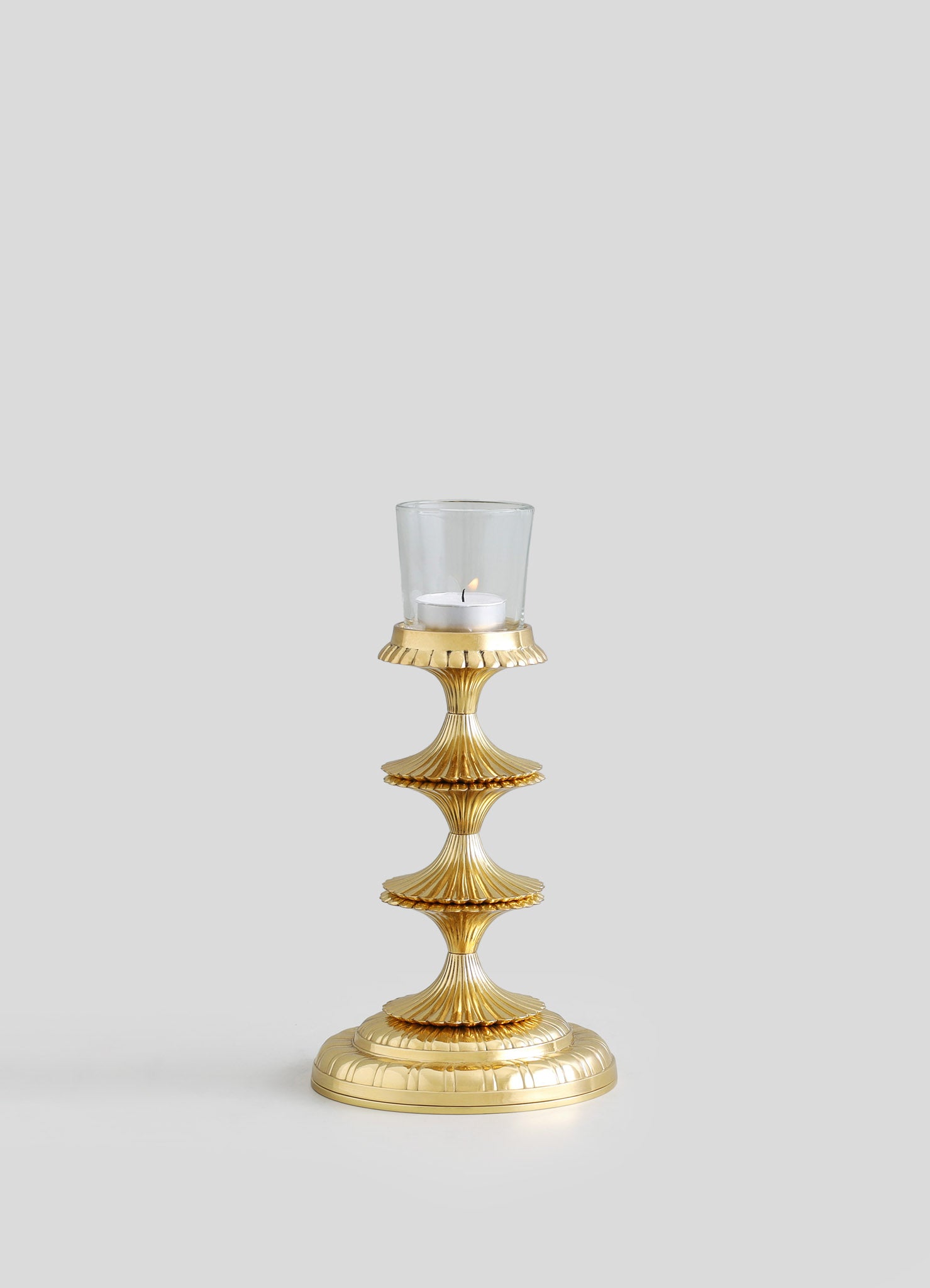 Gold-toned metal candle holder with a ribbed, scalloped design, supporting a glass hurricane with a white tealight candle inside, in a contemporary style.