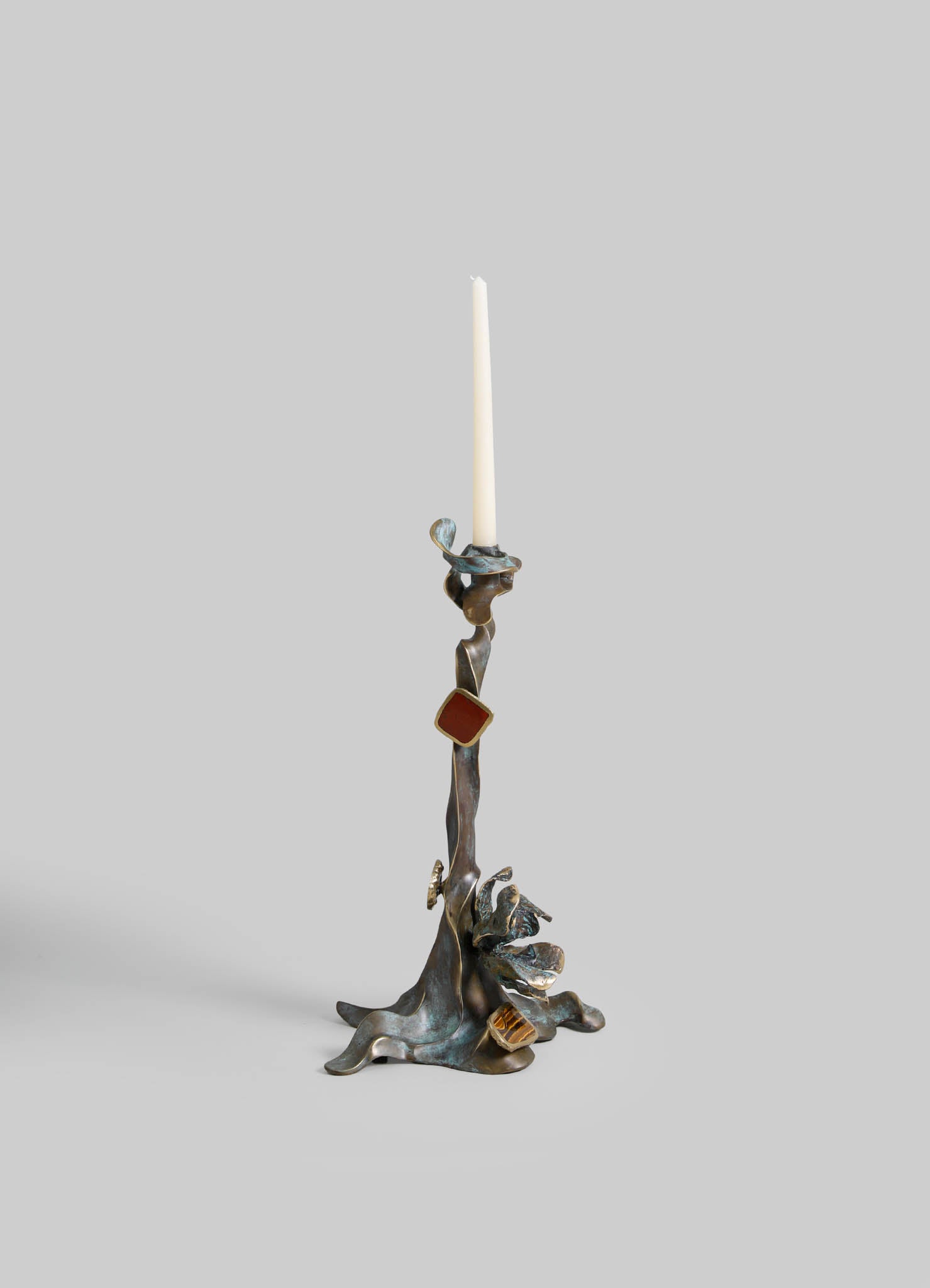 Sculptural candlestick holder with an organic, abstract design in mixed metal tones, featuring irregular shapes and textured surfaces, holding a tall white taper candle.