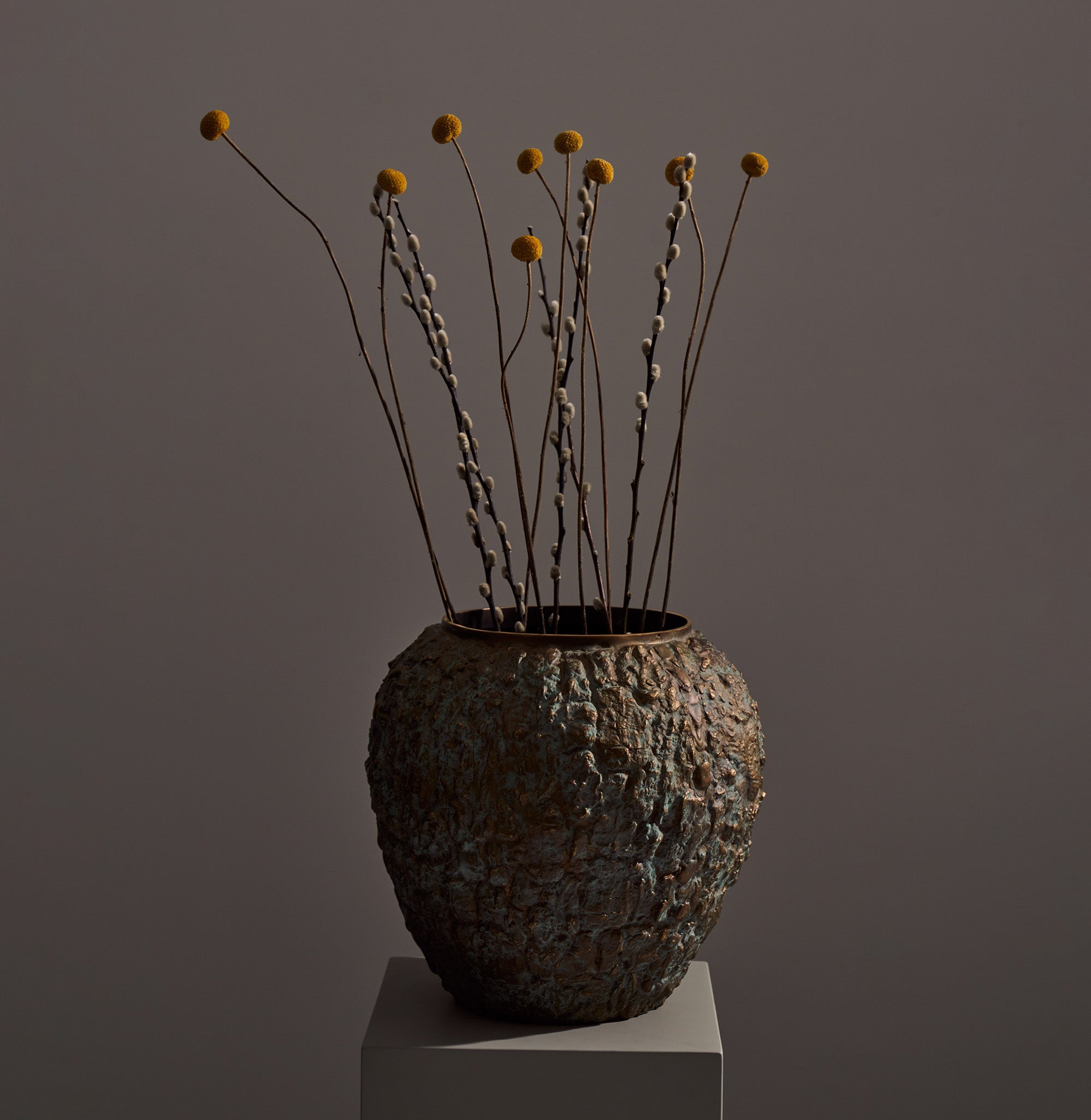 A textured ceramic vase with a weathered, rustic finish in earthy tones, holding dried floral stems with yellow and beige accents.