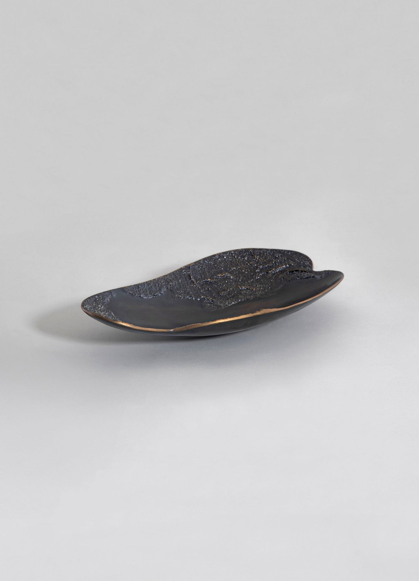 A decorative ceramic dish with a glossy black finish and textured, speckled pattern in gold and bronze tones, featuring an elongated oval shape. The piece has a contemporary design with smooth, curved edges and a subtle metallic accent along the rim.