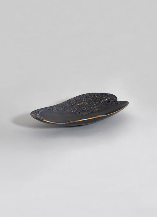 A decorative ceramic dish with a glossy black finish and textured, speckled pattern in gold and bronze tones, featuring an elongated oval shape. The piece has a contemporary design with smooth, curved edges and a subtle metallic accent along the rim.