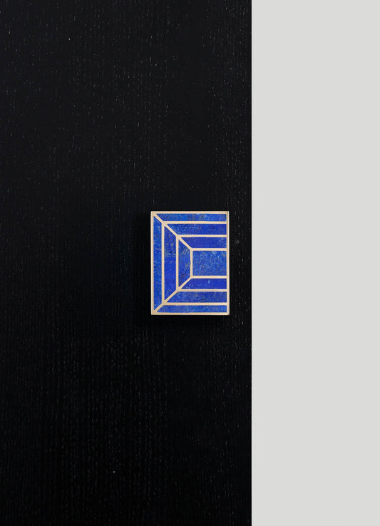 A rectangular ceramic tile with a blue and white geometric pattern featuring concentric lines and angles, displayed on a black textured surface with a light grey background.