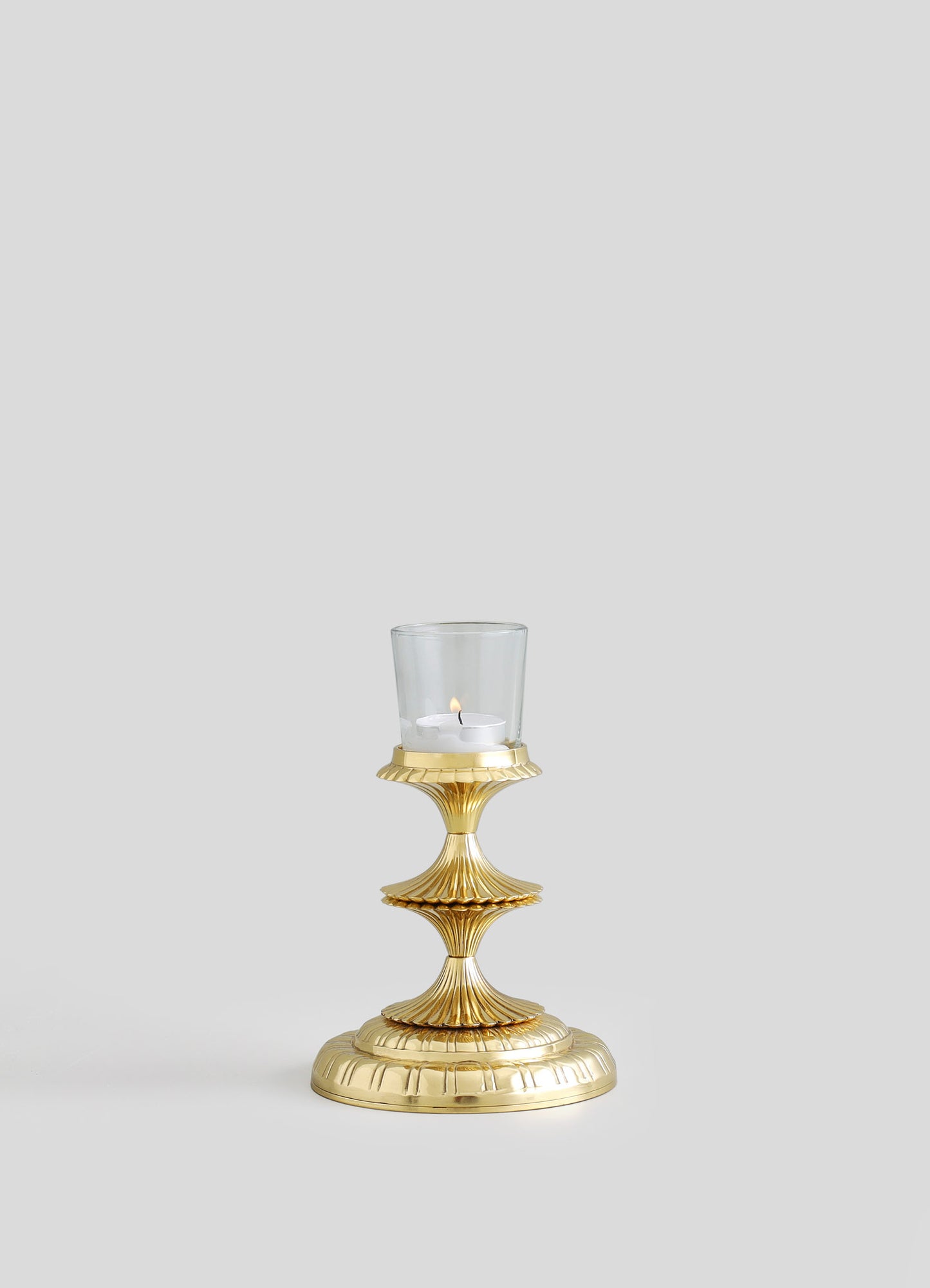 A gold-colored candlestick holder with a scalloped, fluted design, supporting a glass hurricane with a white candle inside.