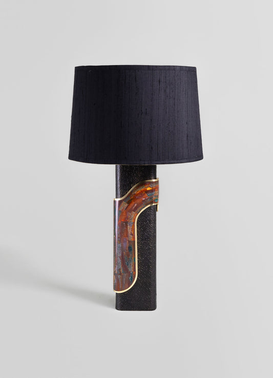 A table lamp with a black textured cylindrical base and a fabric lampshade in solid black. The lamp features a decorative metal accent with a colorful, mosaic-like pattern in warm tones, set against a plain light gray background.