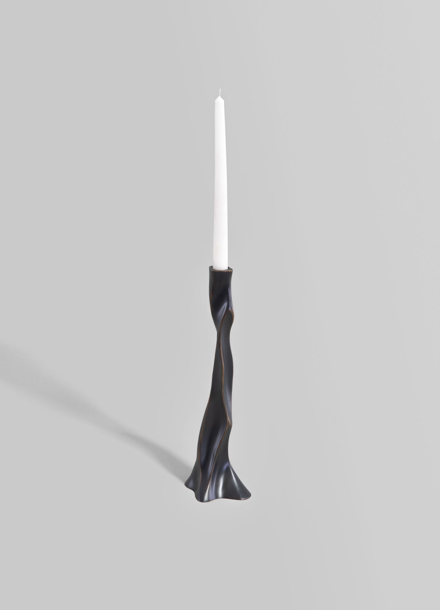 A sculptural candlestick holder with a twisted, organic form in a dark metallic finish, holding a tall white taper candle. The design is modern and contemporary, emphasizing smooth curves and a sleek surface.