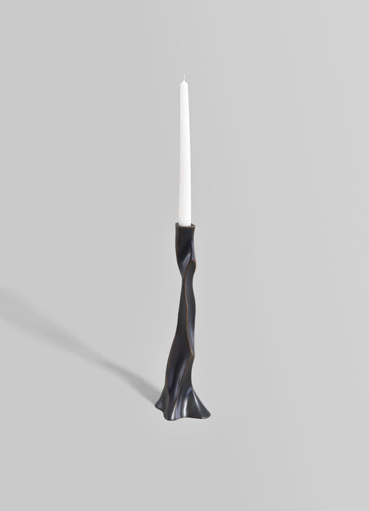A sculptural candlestick holder with a twisted, organic form in a dark metallic finish, holding a tall white taper candle. The design is modern and contemporary, emphasizing smooth curves and a sleek surface.