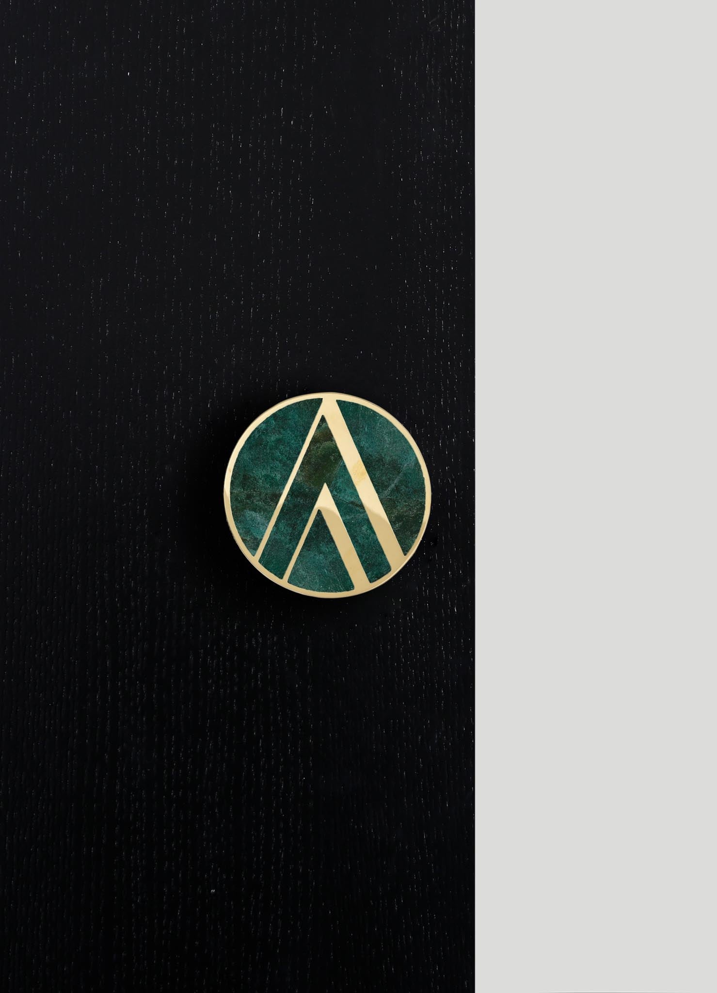 A circular pin with a geometric pattern in gold outline set against a dark fabric background with a textured surface. The pattern features angular lines and shapes, and the pin appears to have an enamel or similar finish.