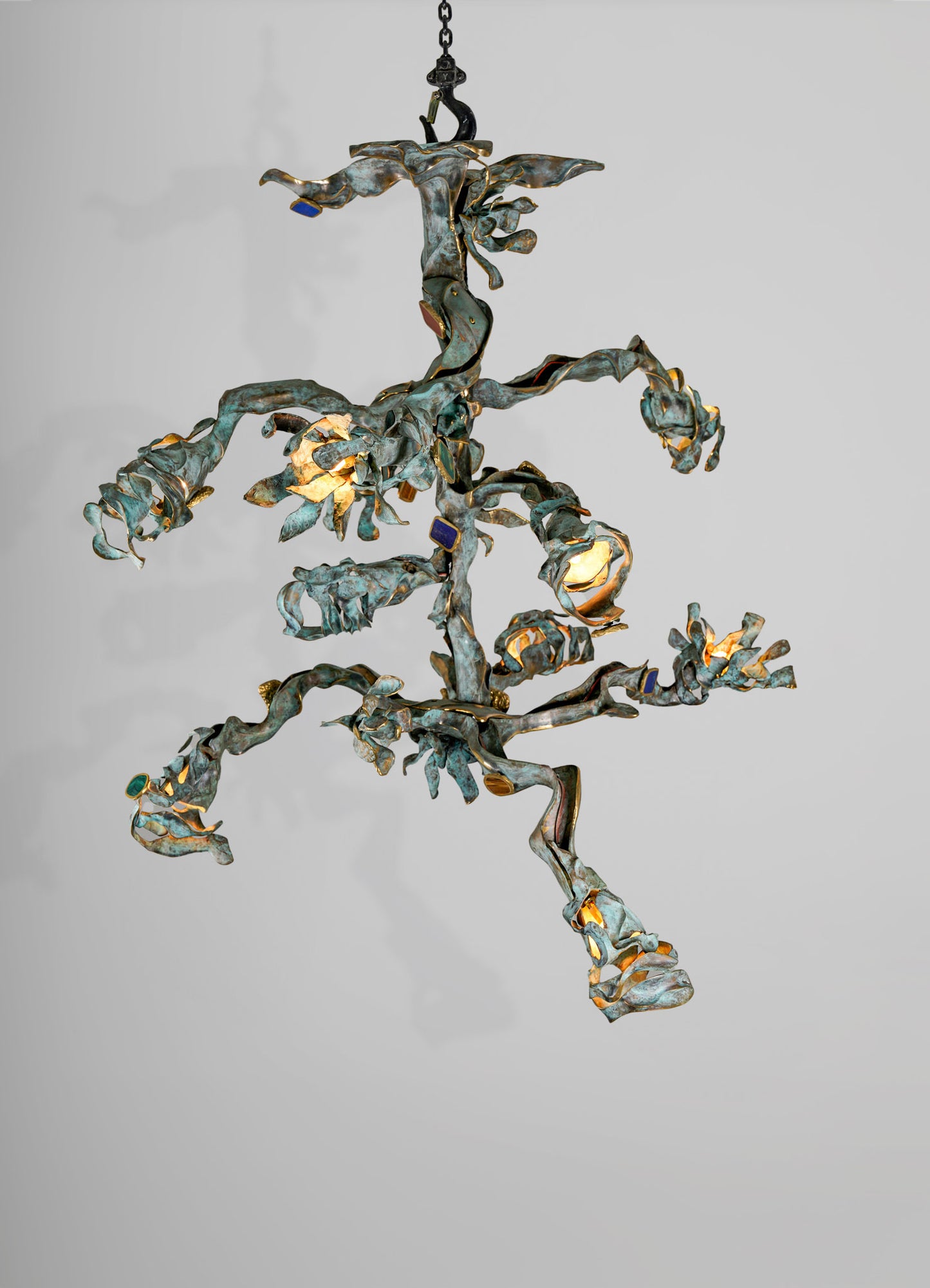 A sculptural pendant light fixture with an organic, leaf-like form crafted from distressed metal in a greenish-blue patina, accented with gold details, and illuminated from within.