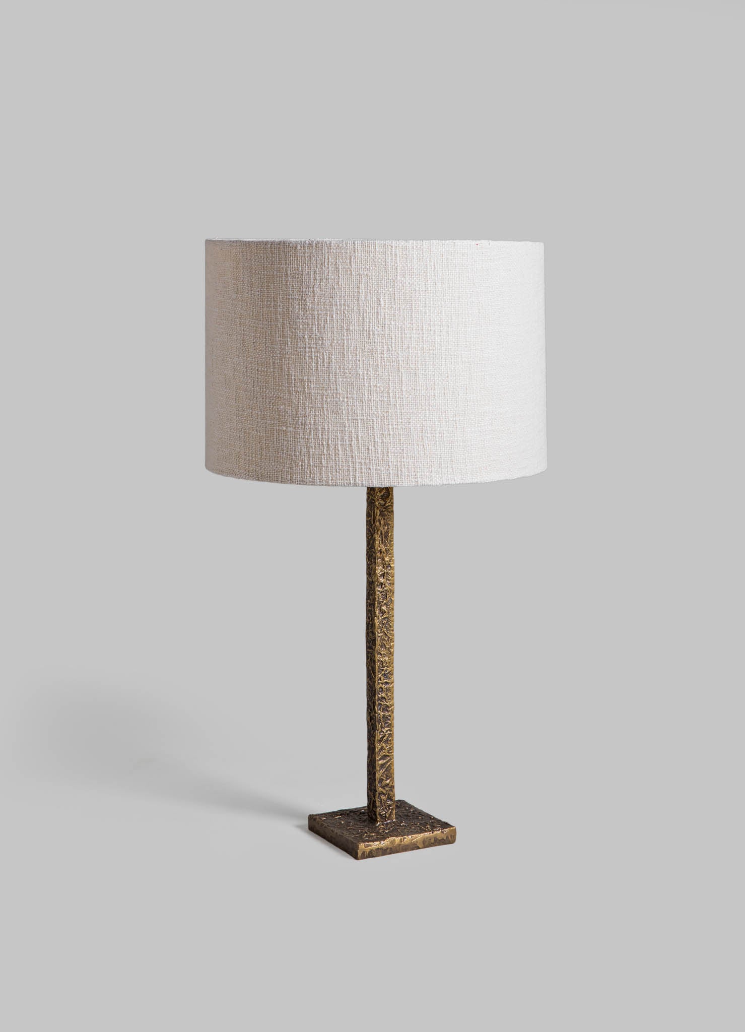 Table lamp with a textured brass base and a cylindrical off-white linen shade, featuring a contemporary design with a neutral color palette.
